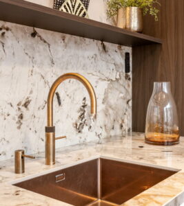 Lichte natuursteen aanrechtblad van keramiek + achterwand, Quooker messing patina en Gold Lanesto spoelbak goud en zeeppomp