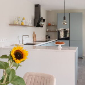 Moderne U-keuken ijsblauw met bar, RVS Quooker en spoelbak, oven met RVS frame, hoogglans wit composiet werkblad en glazen hanglampjes - I-Kook keuken van een trotse klant